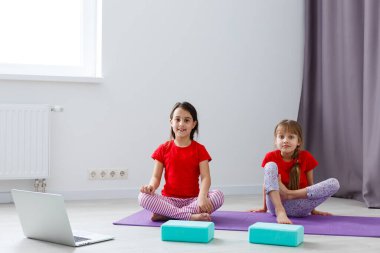 İki küçük kız yoga, esneme, dizüstü bilgisayarda video ile spor yapıyor. Uzaktan eğitim, evde aerobik. Sağlıklı yaşam tarzı, koronavirüs, evde kal. Çocuklar ev karantinası yapıyor.