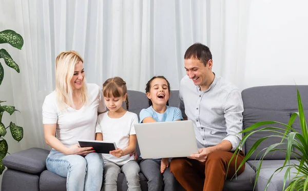 Ung glad avslappnad familj med två barn och par sitter på soffan och tittar på rolig video eller karikatyrer i touchpad — Stockfoto