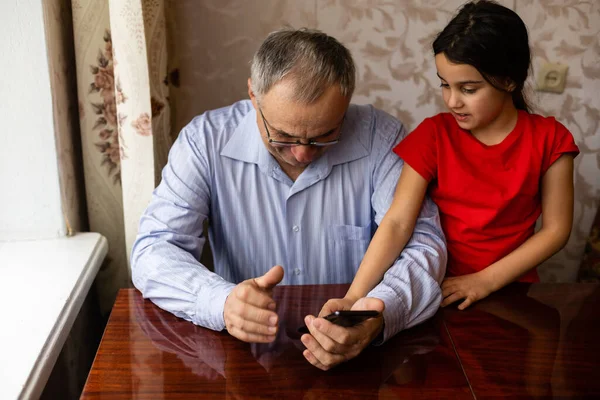 Senior grandfather with child girl granddaughter spending time home together, sitting in living room, using digital mobile phone. Watching videos, playing games, social network — Stock Photo, Image