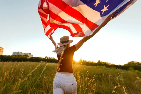 En vacker ung flicka som håller en amerikansk flagga i vinden på ett rågfält. Sommarlandskap mot den blå himlen. Horisontell inriktning. — Stockfoto