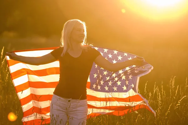 En vacker ung flicka som håller en amerikansk flagga i vinden på ett rågfält. Sommarlandskap mot den blå himlen. Horisontell inriktning. — Stockfoto