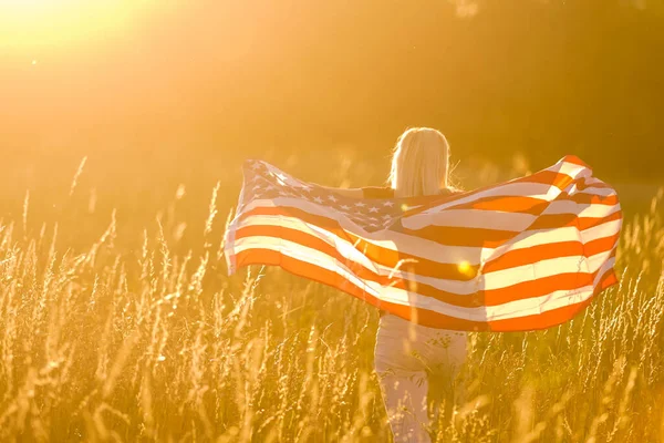 Hermosa joven sosteniendo una bandera americana en el viento en un campo de centeno. Paisaje de verano contra el cielo azul. Orientación horizontal. — Foto de Stock