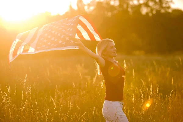 Hermosa joven sosteniendo una bandera americana en el viento en un campo de centeno. Paisaje de verano contra el cielo azul. Orientación horizontal. — Foto de Stock