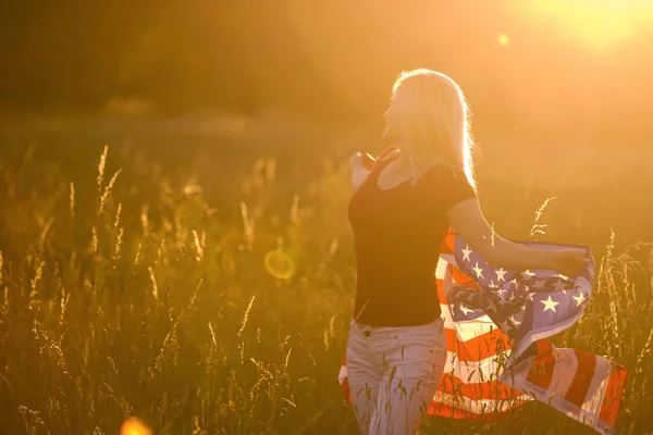 En vacker ung flicka som håller en amerikansk flagga i vinden på ett rågfält. Sommarlandskap mot den blå himlen. Horisontell inriktning. — Stockfoto