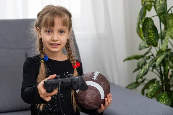 Bambina che tiene una palla da rugby e joystick per giocare ai videogiochi — Foto Stock