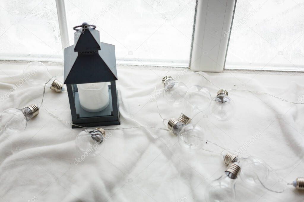 Winter concept. Vintage black lantern with burning candle. Original garland made of lamp bulbs. White snowy winter window on background