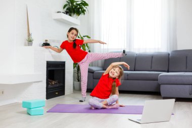 İki küçük kız yoga, esneme, dizüstü bilgisayarda video ile spor yapıyor. Uzaktan eğitim, evde aerobik. Sağlıklı yaşam tarzı, koronavirüs, evde kal. Çocuklar ev karantinası yapıyor.