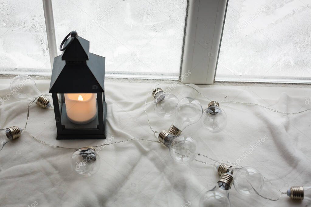 Winter concept. Vintage black lantern with burning candle. Original garland made of lamp bulbs. White snowy winter window on background
