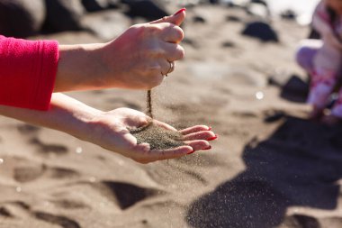 Denize ve kayalara kum döker. Zamanın tükenmesinin ve zaman yönetiminin sembolü. Manikürlü tırnaklar.