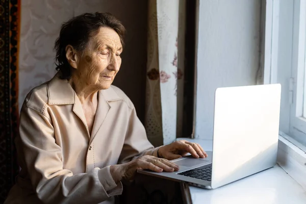 La donna anziana usa il portatile. Sembra molto sorpresa. Navigare in un browser e social media. Un portatile sul tavolo. Utilizzo della tecnologia nella vecchiaia — Foto Stock