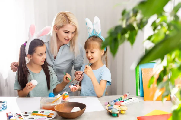 Junge Mutter und ihre beiden kleinen Töchter bemalen zu Hause bunte Ostereier — Stockfoto