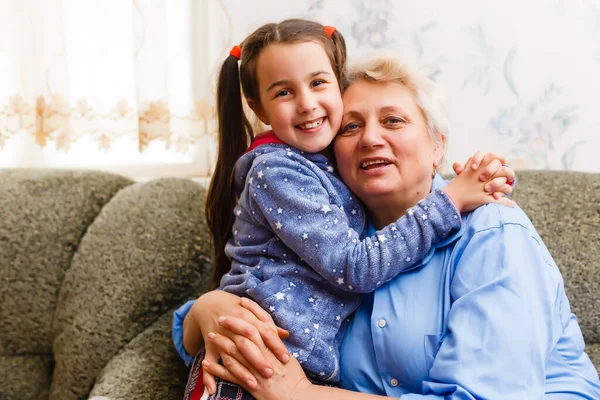 En söt liten flicka och hennes mormor umgås hemma. Ha kul, kramas och le när du sitter på soffan. — Stockfoto