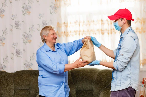 Joven voluntario con máscara le da a una anciana cajas con comida cerca de su casa. Hijo el hombre ayuda a una madre anciana soltera. Apoyo familiar, cariño. Cuarentena, aislada. Coronavirus covid-19. Donación — Foto de Stock