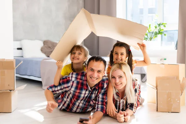 Familia caucásica, hombre, mujer y dos niñas se sientan en el suelo, desempacan cajas y sonríen en la nueva casa. Detrás de ellos cajas móviles. —  Fotos de Stock