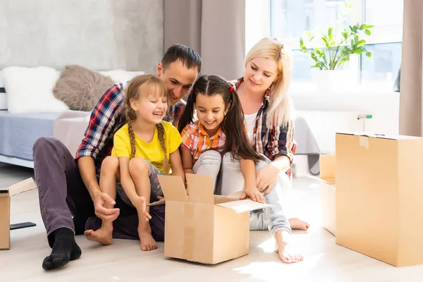Blanke familie, man, vrouw en twee meisjes zitten op de vloer, pakken dozen uit en lachen in een nieuw huis. Achter hen verplaatsen dozen. — Stockfoto