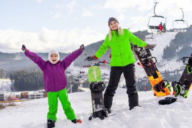 Snowboard yapan anne ve kızı karda oynuyorlar.