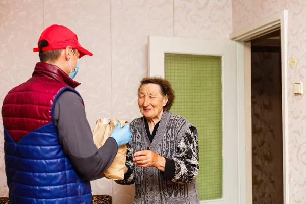 Jonge mannelijke vrijwilliger in masker geeft een oudere vrouw dozen met voedsel in de buurt van haar huis. Zoon man helpt een alleenstaande oudere moeder. Familie steun, zorg. In quarantaine, geïsoleerd. Coronavirus covid-19. Donatie — Stockfoto