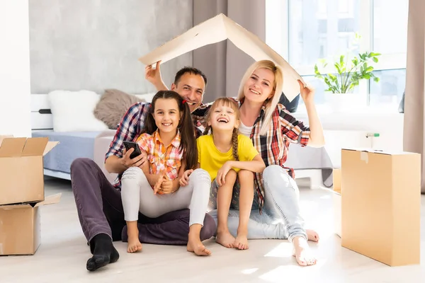 Blanke familie, man, vrouw en twee meisjes zitten op de vloer, pakken dozen uit en lachen in een nieuw huis. Achter hen verplaatsen dozen. — Stockfoto