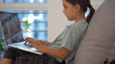 Okul çağındaki küçük kız evde okuyor. Bir kız dizüstü bilgisayar ekranından video dersi izler ve görevleri not defterine yazar..