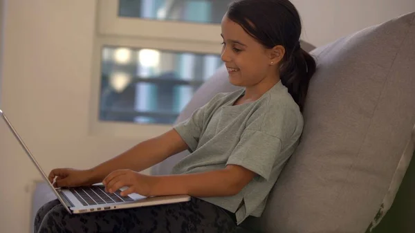 Holčička ve školním věku se učí doma. Dívka se dívá na video lekci na notebooku monitoru a píše úkoly do notebooku. — Stock fotografie
