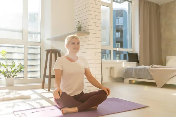 Hora del yoga. Atractiva joven mujer haciendo ejercicio y sentada en posición de loto de yoga mientras descansa en casa — Foto de Stock