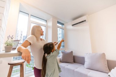 Havalandırma altında mutlu bir aile, oturma odasındaki uzaktan kumandalı bir anne kız için konfor ısısını, modern evdeki iklim sistemini ayarlıyor.