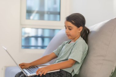 Okul çağındaki küçük kız evde okuyor. Bir kız dizüstü bilgisayar ekranından video dersi izler ve görevleri not defterine yazar..