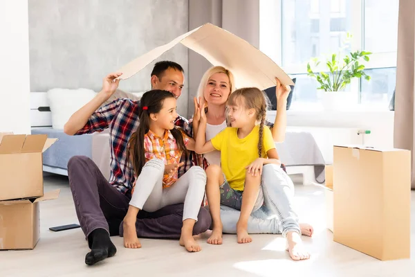 Heyecanlı genç aile, taşınma günü karton kutularını açarken eğleniyor. Mutlu ebeveynler, küçük kızlarıyla oynuyor. Karton paketten atlıyorlar.