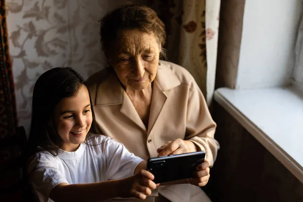 A bisavó senta-se com a bisneta e olha para o smartphone. Avó e criança tirar uma selfie em um smartphone. Avó com sua neta olhar para o telefone. — Fotografia de Stock