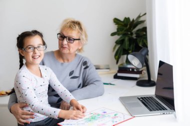 Yetişkin bir büyükanne, çocuğun ödevlerine evde yardım ediyor. Tatmin olmuş yaşlı büyükanne torununa oturma odasında ders çalışmasında yardım ediyor. Küçük kız yanında öğretmeniyle birlikte deftere yazı yazıyor.