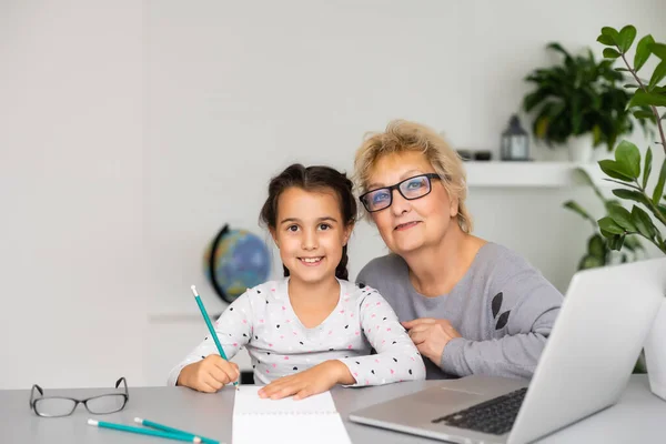 La nonna matura aiuta il bambino con i compiti a casa. Soddisfatto vecchia nonna aiutare la nipote a studiare in soggiorno. Ragazzina che scrive sul taccuino con l'insegnante anziana seduta accanto a lei — Foto Stock