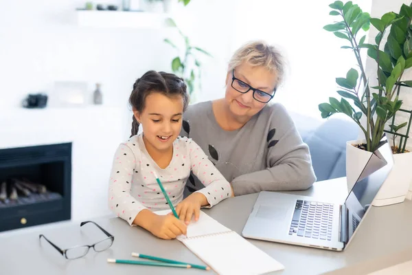 Starší babička pomáhá dítěti s domácími úkoly. Spokojen stará babička pomáhá své vnučce studovat v obýváku. Malá dívka psaní na notebook s senior učitel sedí vedle ní — Stock fotografie