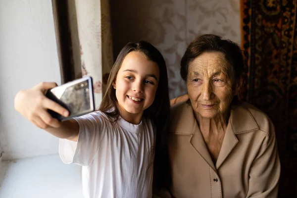 La bisabuela se sienta con su bisnieta y mira al teléfono inteligente. La abuela y el niño se toman una selfie en un teléfono inteligente. Abuela con su nieta mirar en el teléfono. — Foto de Stock