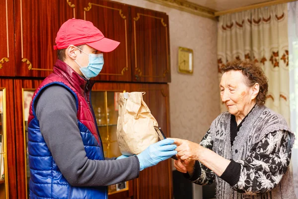 Joven voluntario con máscara le da a una anciana cajas con comida cerca de su casa. Hijo el hombre ayuda a una madre anciana soltera. Apoyo familiar, cariño. Cuarentena, aislada. Coronavirus covid-19. Donación —  Fotos de Stock