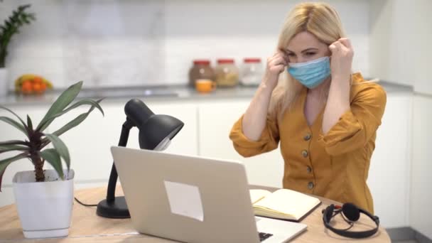 Mujer en máscara protectora funciona de forma remota en el ordenador portátil en casa en la cocina, la mujer se quita la máscara — Vídeos de Stock