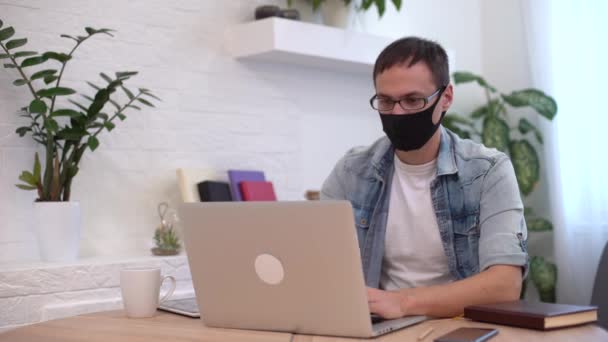 De jonge blanke man blijft thuis en draagt een beschermend masker met laptop en internet. Gezellig thuiskantoor, werkplek coronavirus pandemie, covid 19 quarantaine. Afstandswerk, freelancer — Stockvideo