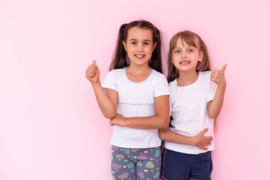 Çocuklar beğeniyor. İki sevimli mutlu küçük kız başparmaklarını bir araya getirip kameraya gülümsüyor, mükemmel geri bildirim, iyi iş. Pembe arka planda kapalı stüdyo çekimleri