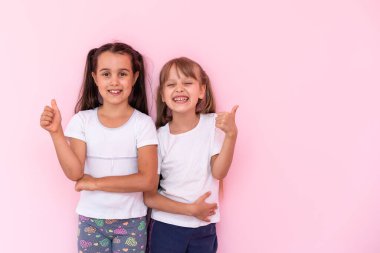 Çocuklar beğeniyor. İki sevimli mutlu küçük kız başparmaklarını bir araya getirip kameraya gülümsüyor, mükemmel geri bildirim, iyi iş. Pembe arka planda kapalı stüdyo çekimleri