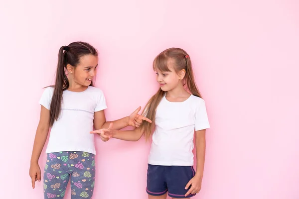Niñas niños asombrados mostrando camiseta blanca, aislado sobre fondo rosa. Concepto de estilo de vida infantil. Prepara espacio para copias. Señalar el dedo índice a un lado — Foto de Stock