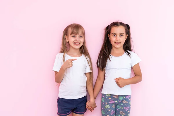 Niñas niños asombrados mostrando camiseta blanca, aislado sobre fondo rosa. Concepto de estilo de vida infantil. Prepara espacio para copias. Señalar el dedo índice a un lado — Foto de Stock
