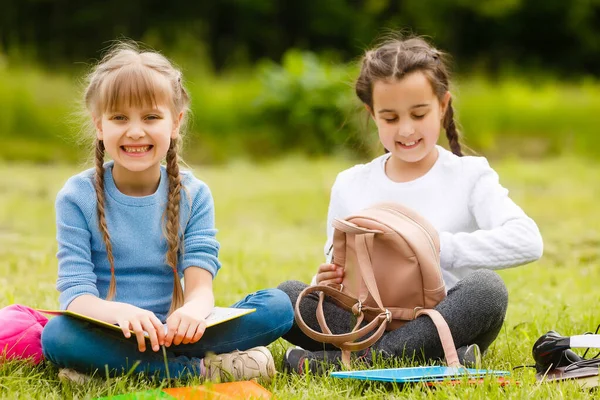 Két csinos iskoláslány ül kint könyvekkel a parkban. Iskoláslányoknak vagy diákoknak tanítják a természetet.. — Stock Fotó