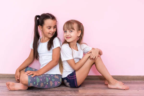 Dos niñitas sentadas en el suelo y mirando hacia arriba. Aislado sobre un fondo rosa — Foto de Stock