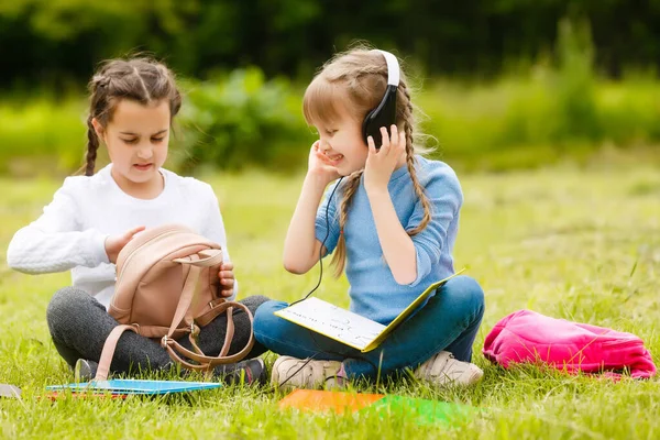 Zwei lustige Schulmädchen sitzen auf dem Rasen und lesen Bücher. Mädchen, Freundinnen, Schwestern werden Lektionen in der Natur erteilt. — Stockfoto