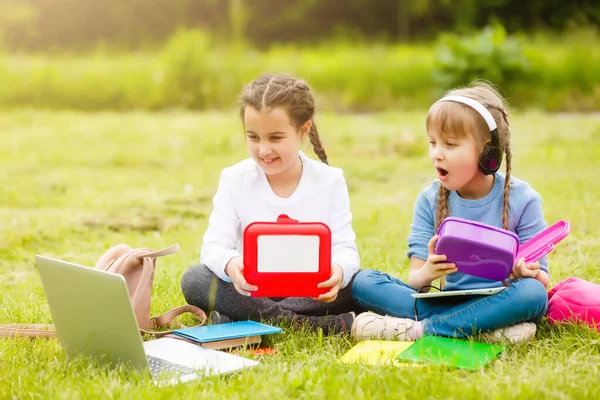 Žáci základní školy s krabicemi od oběda v rukou. Holky s batohy jedí na trávníku. Začátek lekcí. První den podzimu. — Stock fotografie