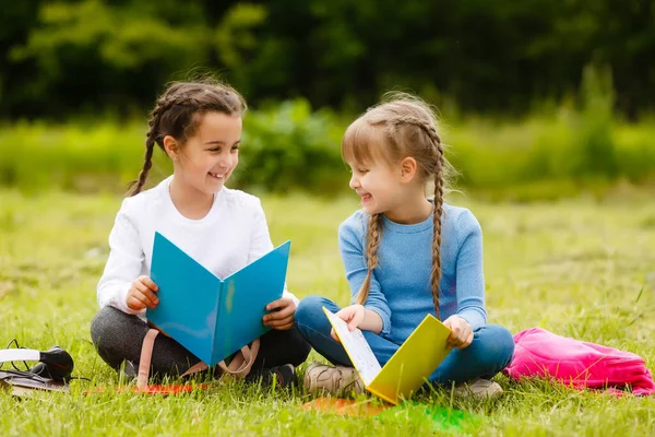 Två söta multikulturella skolflickor sitter på gräsmattan under träd och läser bok tillsammans — Stockfoto