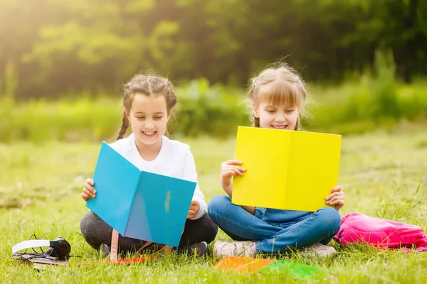 Zwei süße multikulturelle Schulmädchen sitzen auf dem Rasen unter einem Baum und lesen zusammen ein Buch — Stockfoto