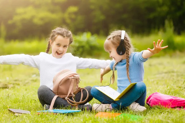Twee leuke multiculturele schoolmeisjes zitten op het gazon onder de boom en samen lezen boek — Stockfoto