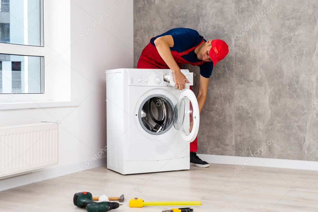Plumber in overalls with tools is repairing a washing machine in the house