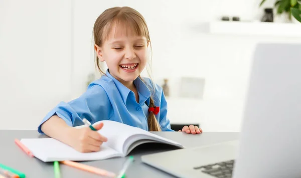 Az okos kislány elvégzi a házi feladatot a nappaliban. Az íróasztalánál ül, tollal ír a tankönyvében, és laptopot használ.. — Stock Fotó