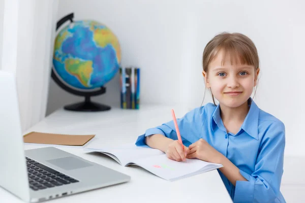 Έξυπνο κοριτσάκι κάνει τις εργασίες στο σαλόνι της. Shes κάθεται στο γραφείο της γράφει με ένα στυλό στα βιβλία της και χρήσεις Laptop. — Φωτογραφία Αρχείου